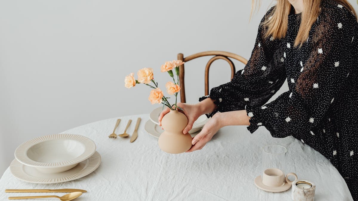 A minimalist table setting featuring a woman arranging flowers in a modern vase.